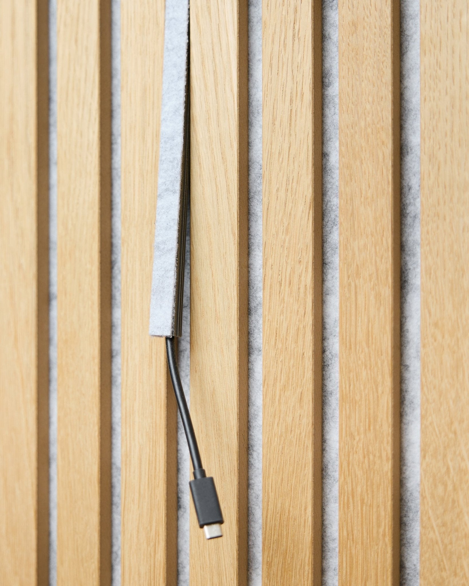 En black USB-C-kabel går gjennom et grey WoodUpp Dupps Pop-on Cable Cover mellom vertical akustiske trespiler med grey filt på baksiden.